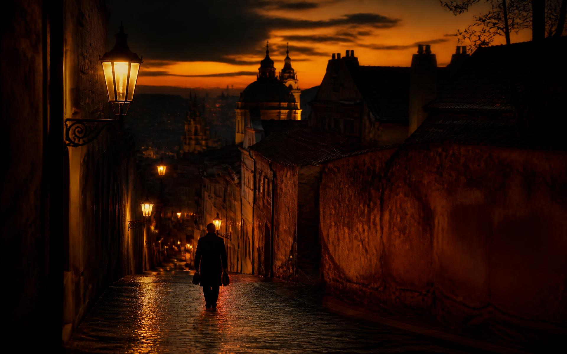 A lone traveller on a lamplit old-world cobblestone street at golden dusk